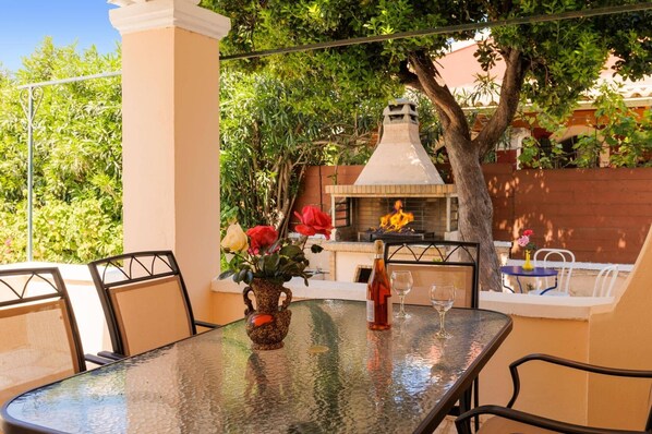 Eine Terrasse im Freien mit einem Glastisch, Stühlen, einer Vase mit Blumen, einer Flasche und einem Glas. Im Hintergrund steht ein Grill und ein Baum.
