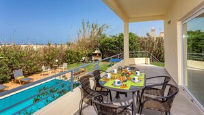 A balcony with a pool, dining table, and chairs, overlooking a garden and cityscape.