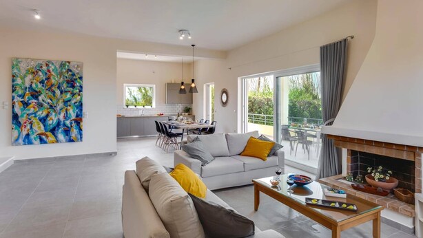A spacious living area with a sofa, coffee table, and a fireplace. There is a dining area with a table and chairs, and a kitchen in the background.