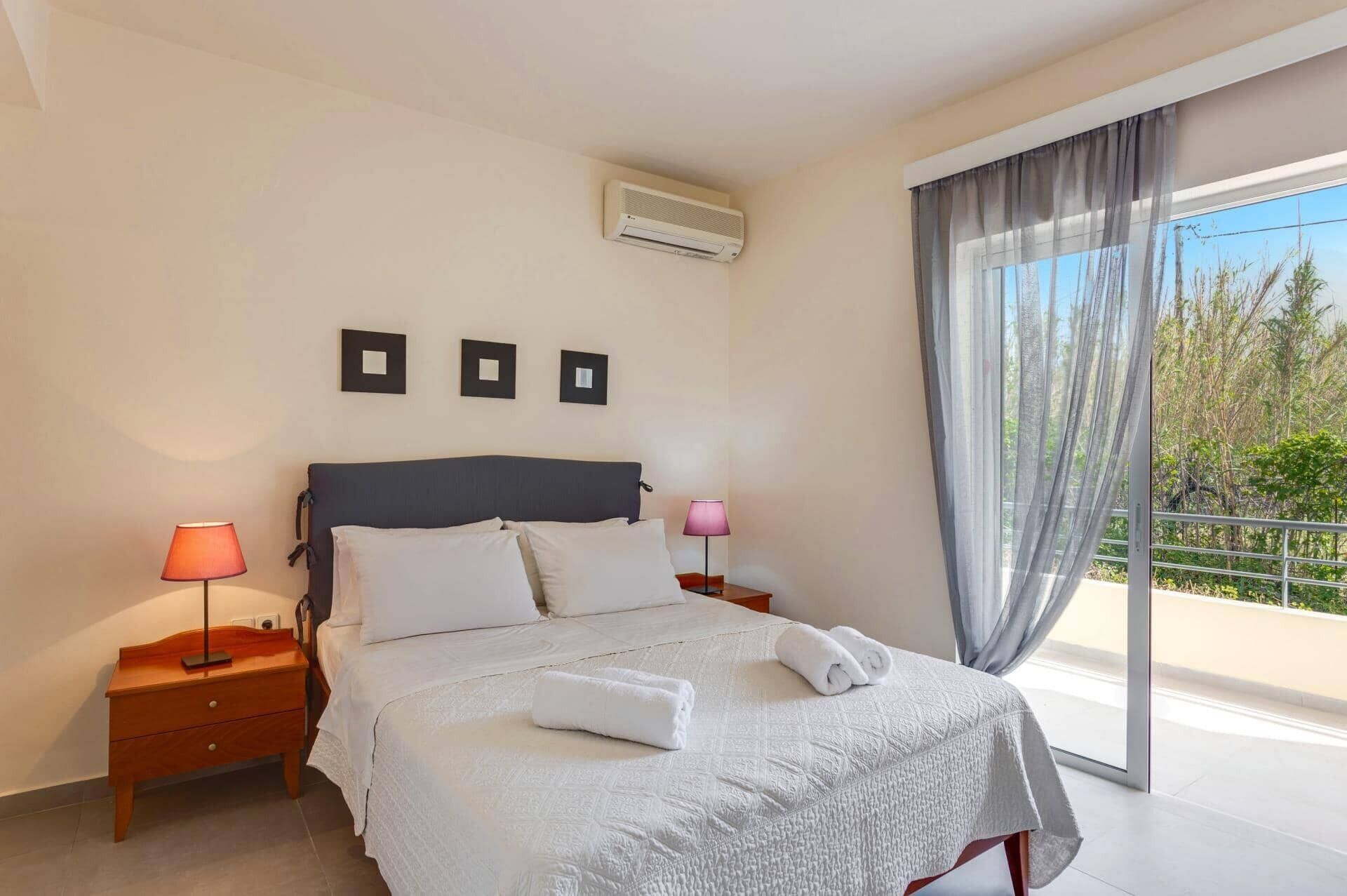 A bedroom with a bed, two bedside tables, a nightstand, a window with curtains, and an air conditioning unit.