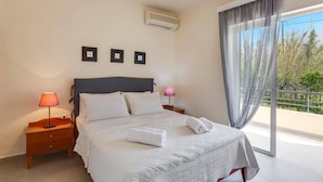 A bedroom with a bed, two bedside tables, a nightstand, a window with curtains, and an air conditioning unit.