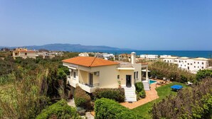 A coastal residential area with a modern house featuring a swimming pool, a view of the sea, and a hilly landscape in the background.