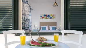 Mesa de café da manhã com fatias de melancia, um copo de suco de laranja e uma tigela de granola. Há uma cama com almofadas azuis e uma pintura colorida ao fundo.