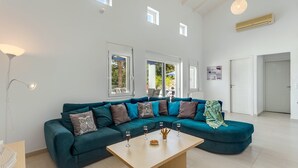 A spacious living room with a teal sectional sofa, a wooden coffee table, and a large window providing natural light. - Villa Almyrida View by Villa Plus (Apokoronos)