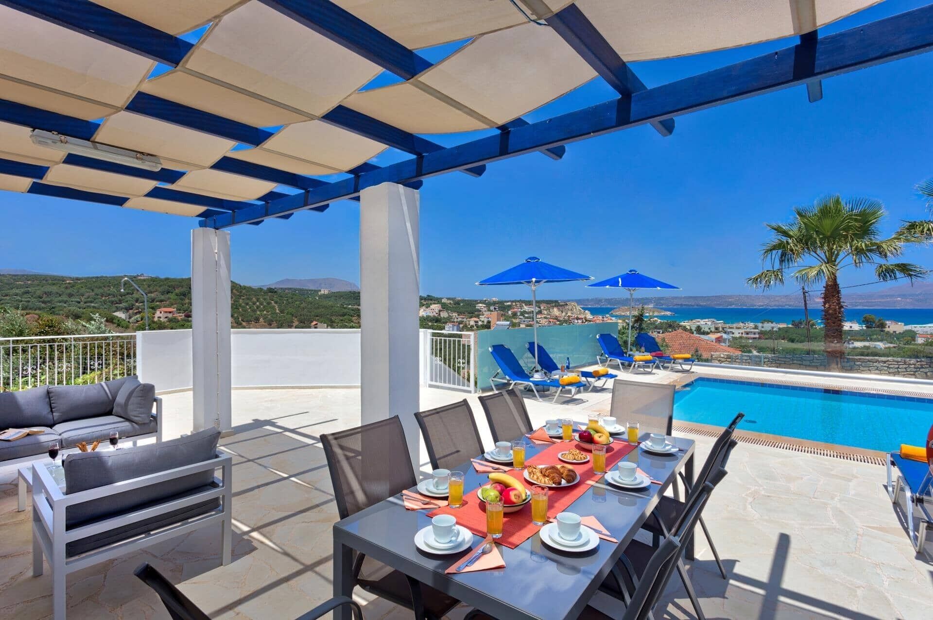 A terrace with a pool, dining table set with plates, glasses, and fruit, and blue umbrellas.