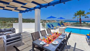 A terrace with a pool, dining table set with plates, glasses, and fruit, and blue umbrellas. - Villa Almyrida View by Villa Plus (Apokoronos)