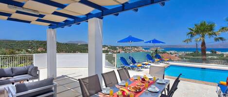 A terrace with a pool, dining table set with plates, glasses, and fruit, and blue umbrellas.