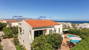 En villa med ett rött tegeltak, ett poolområde med solstolar och utsikt över havet.