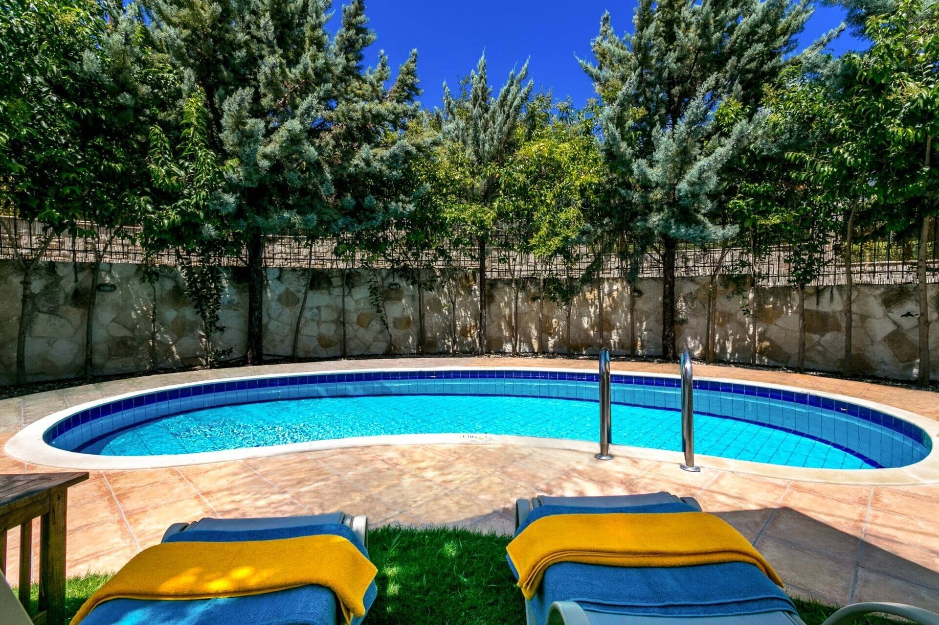 A swimming pool with two lounge chairs, surrounded by a stone wall and trees.