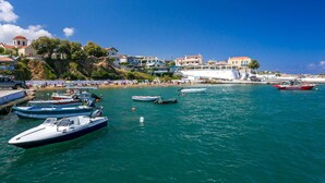 A coastal town with boats docked along the shore, a beach with people, and buildings with balconies overlooking the sea. - Villa Areva by Villa Plus (Panormos)