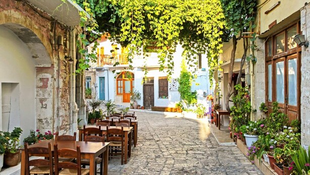 A cobblestone street with wooden tables and chairs, potted plants, and buildings with balconies and windows. - Villa Areva by Villa Plus (Panormos)