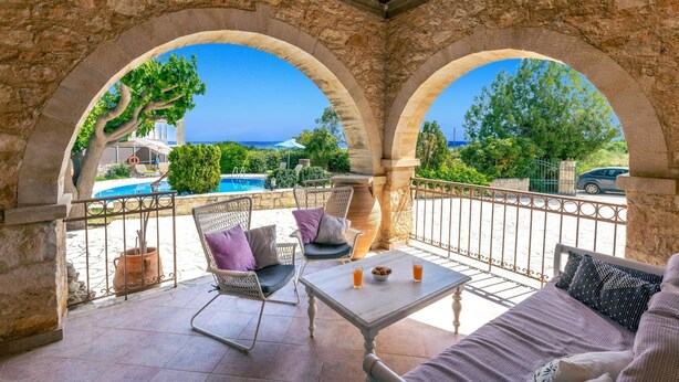 Un espacio de estar al aire libre techado con vista a la piscina, arcos de piedra y una mesa con bebidas.