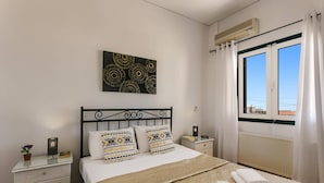 A bedroom with a bed, bedside tables, a wall art piece, a window with curtains, and a radiator. - Villa Capri Crete by Villa Plus (Chersonisos)