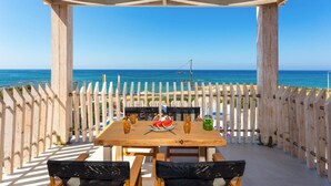 Een houten tafel met stoelen en een tafelkleed, geplaatst op een terras met uitzicht op een strand en een helder zicht op de oceaan, met een boot in de verte.