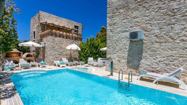 Una piscina con sillas de descanso y sombrillas, rodeada de muros de piedra y vegetación.