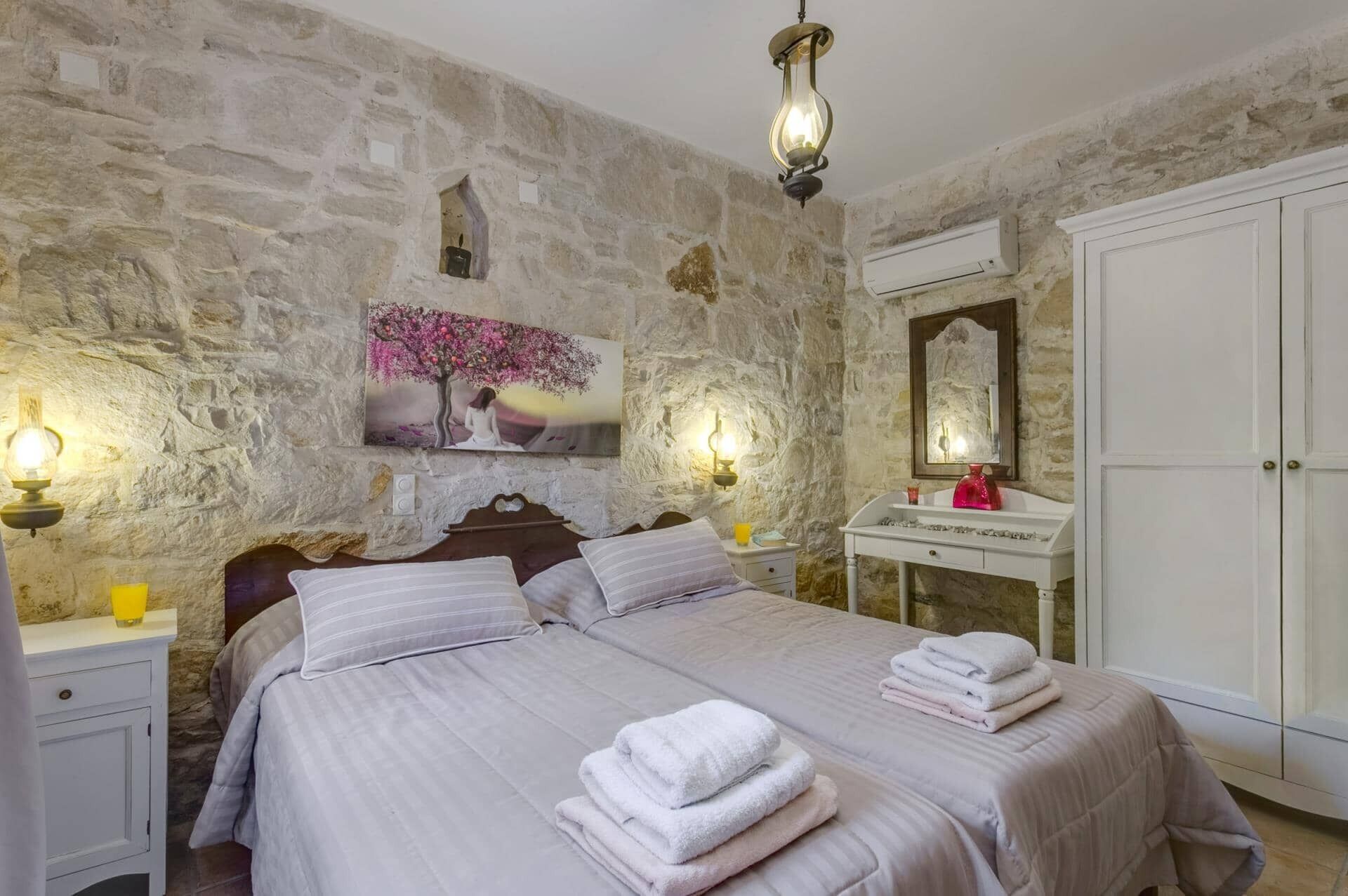 Une chambre avec des murs en pierre, un lit recouvert de draps blancs, une table de chevet, une commode, un miroir et un luminaire fixé au mur.