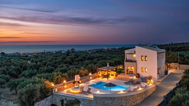 A modern villa with a pool, outdoor seating, and a view of the sea at sunset.