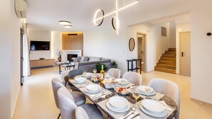 A modern dining area with a long table set for six, a sofa, and a TV in the background.