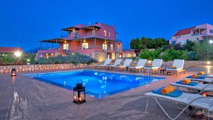 A poolside area with lounge chairs and a lantern, surrounded by a stone wall and a large building with multiple balconies. - Villa Horizon by Villa Plus (Kampani)