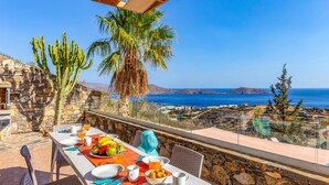 A dining table set with plates, cutlery, and a fruit bowl, overlooking a coastal landscape with cacti and palm trees. - Villa Elounda Pelagos by Villa Plus (Agios Nikolaos Municipality)