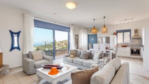A modern living room with a large window, a dining area, and a kitchen. - Villa Elounda Pelagos by Villa Plus (Agios Nikolaos Municipality)