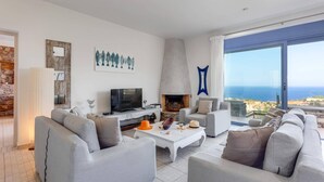 A modern living room with a fireplace, comfortable seating, and a large window offering a view of the sea. - Villa Elounda Pelagos by Villa Plus (Agios Nikolaos Municipality)