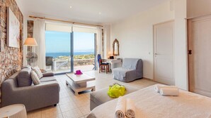 A modern hotel room with a balcony, a grey sofa, a small table, a chair, and a bed with towels. - Villa Elounda Pelagos by Villa Plus (Agios Nikolaos Municipality)