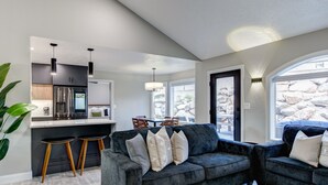 Ein modernes Wohnzimmer mit einem dunkelgrauen Sofa, zwei Holzhockern und einer Küchenzeile mit Edelstahlgeräten.