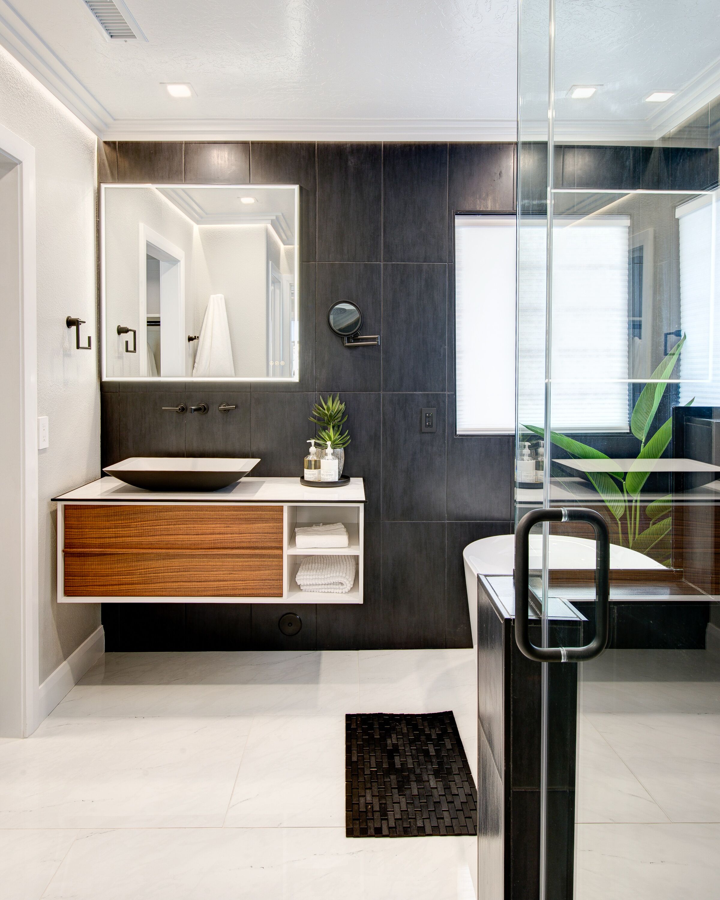 A modern bathroom with a large mirror, dark tiled walls, a wooden vanity, and a glass shower enclosure.