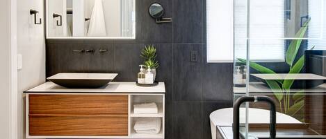 A modern bathroom with a large mirror, dark tiled walls, a wooden vanity, and a glass shower enclosure.