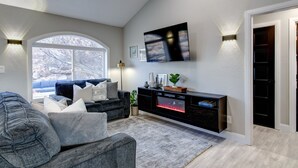Ein modernes Wohnzimmer mit großem Fenster, Kamin, Flachbildfernseher und einem dunkelgrauen Sofa.