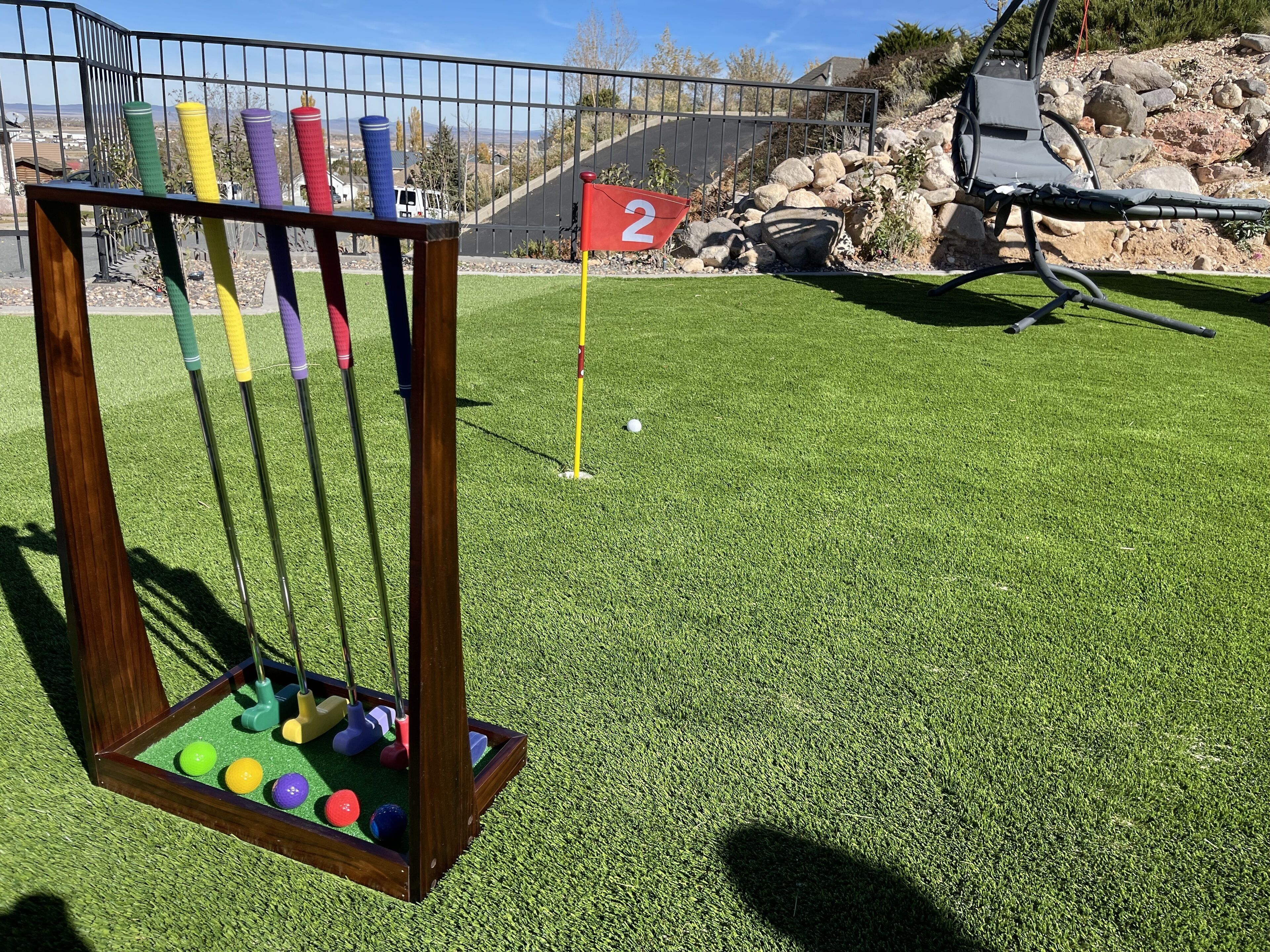 A mini-golf setup with a wooden rack holding colorful golf balls and sticks, a red flag marking the hole, and a green artificial turf.