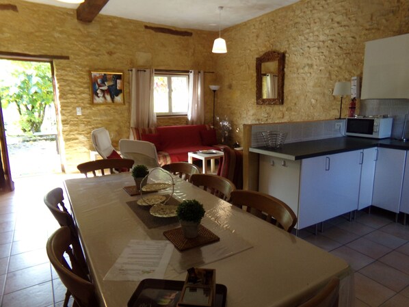 A dining area with a table set for four, chairs, a kitchen with a microwave, and a stone wall. - Pretty gite in the heart of the Dordogne valley sleeping 8 max (7adults) (Nadaillac de rouge)