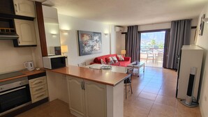 Cozinha compacta com ilha central, área de refeições com sofá vermelho, e vista para o exterior por uma porta de correr.