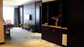 A hotel room with a large dark wooden wardrobe, a flat-screen TV mounted on the wall, a small table with a vase of flowers, and a seating area with chairs and a bench. - Lisan Garden Hotel (Ruichang)