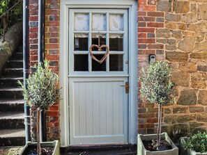 A white door with a heart-shaped window decoration, flanked by two potted plants, leading to a stone wall and a staircase.