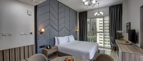 Habitación de hotel moderna con una cama grande, un escritorio con una computadora, una silla, una mesita con un arreglo floral y vistas de la ciudad a través de grandes ventanales.