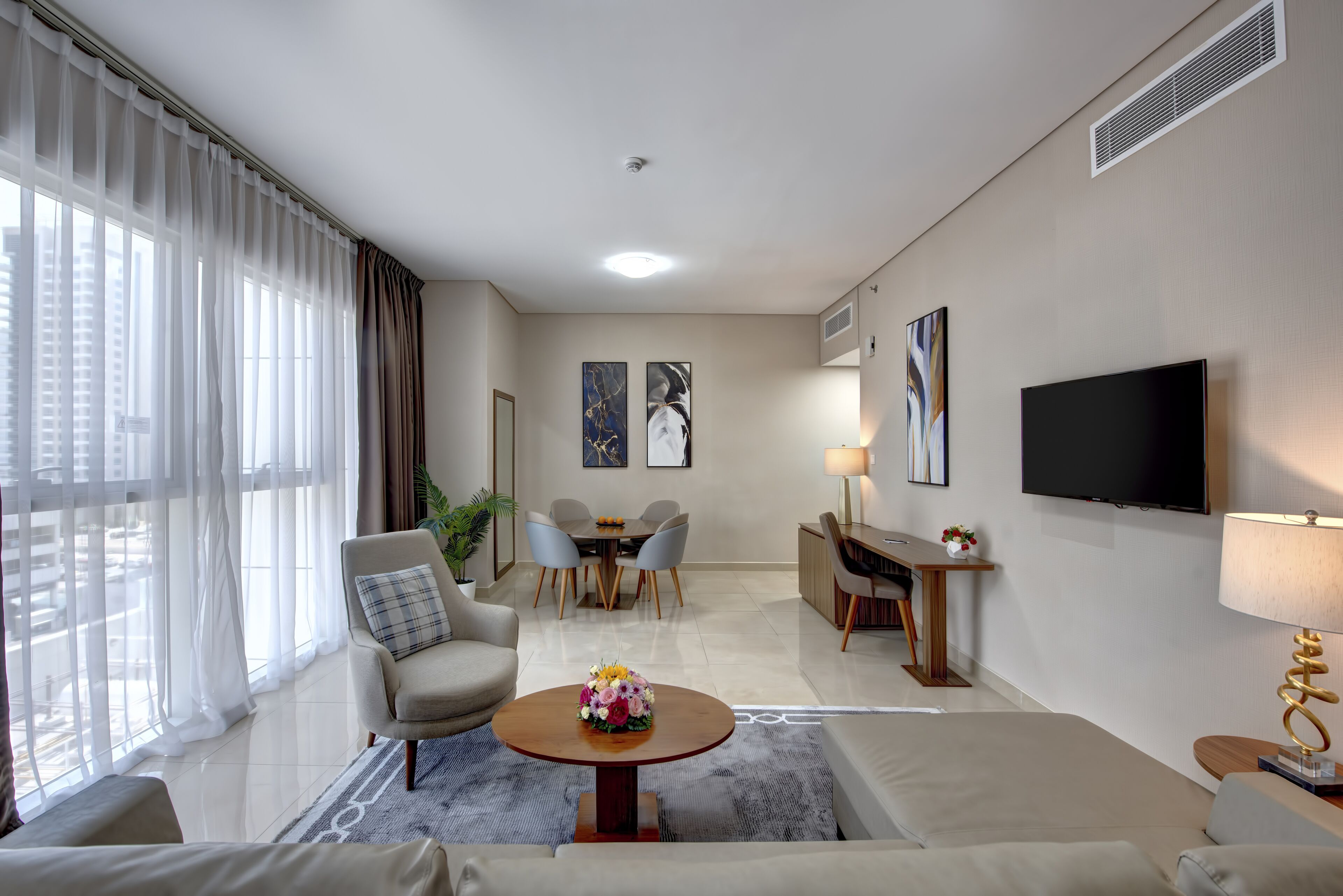 Ein modernes Wohnzimmer mit einer Couch, Sesseln, einem Couchtisch mit Blumen, einem Essbereich mit Stühlen, einem Fernseher und Kunstwerken an den Wänden.