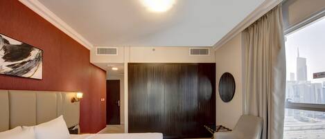 A hotel room with a large bed, a seating area with a chair and a small table, a view of the city through a window, and a door leading to another room.