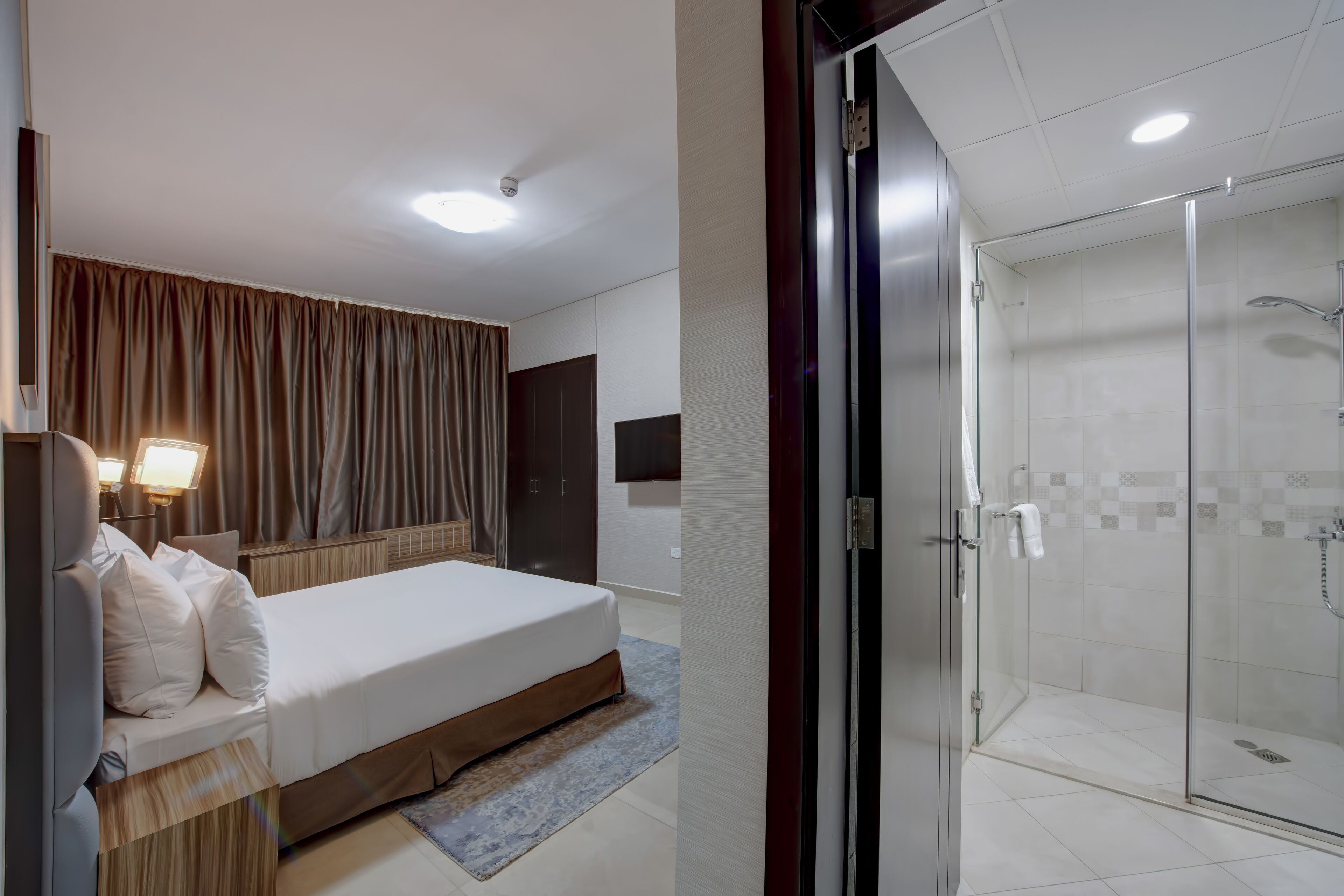A hotel room with a bed, a nightstand, a lamp, and a glass-enclosed shower area.