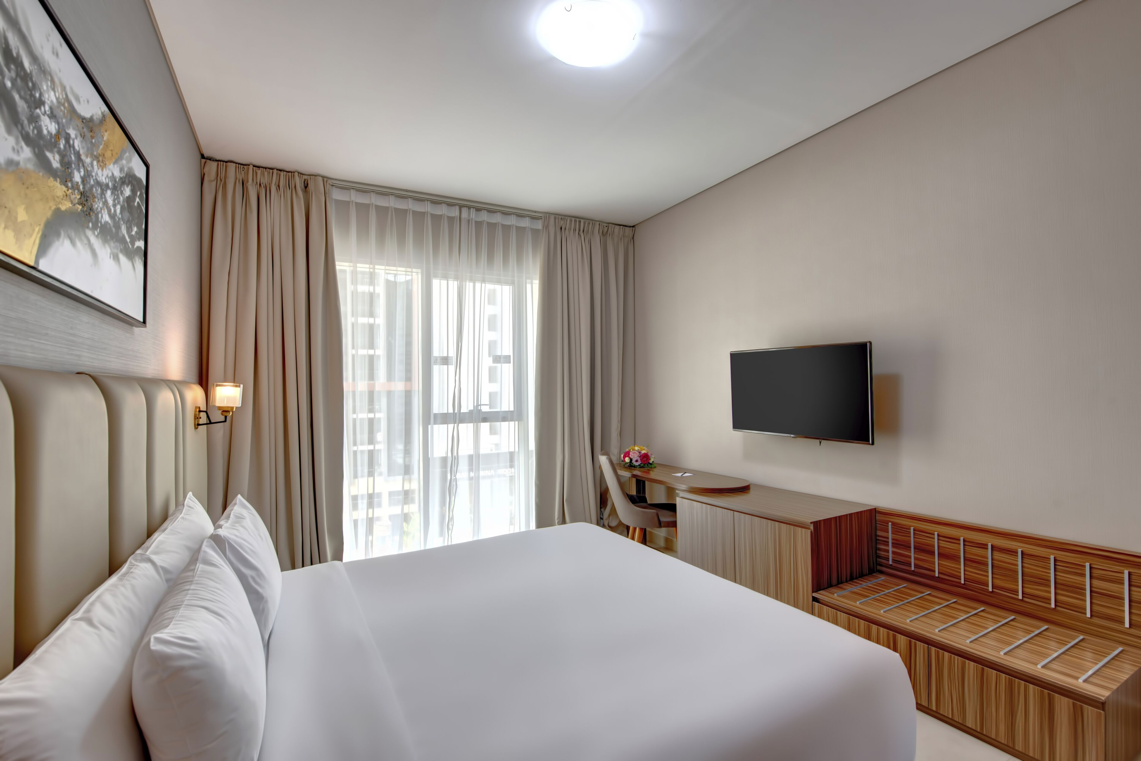 Ein Hotelzimmer mit einem großen Bett, einem Schreibtisch mit Stuhl, einem an der Wand befestigten Fernseher und einem Fenster mit Vorhängen.
