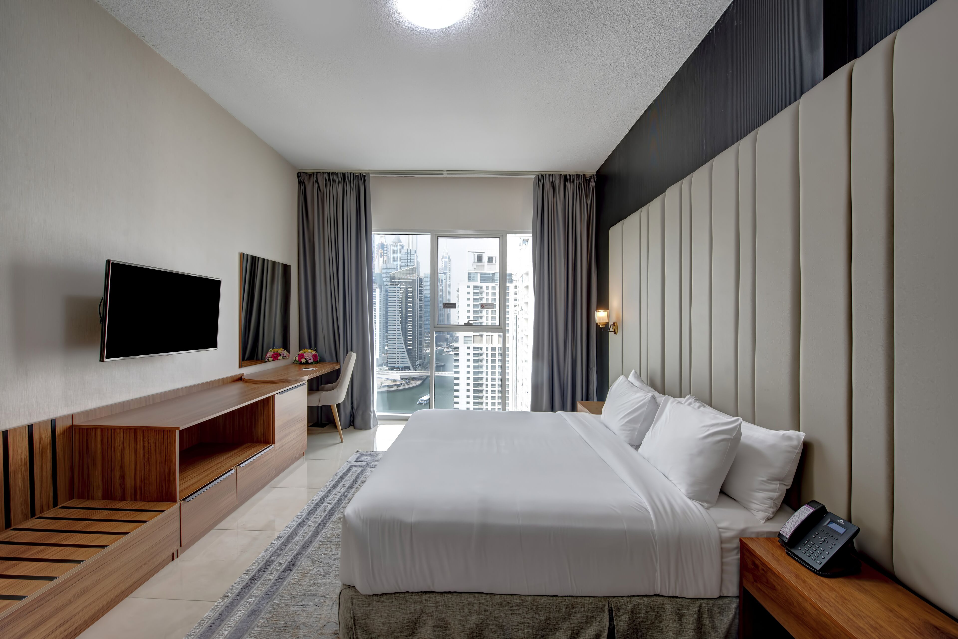 Quarto de hotel moderno com uma cama grande, televisão de tela plana, mesa de console de madeira e vista para a paisagem urbana.