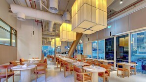 A modern restaurant with round tables and orange chairs, large windows, and a view of the city. - Regency Luxury Suites (Dubai)