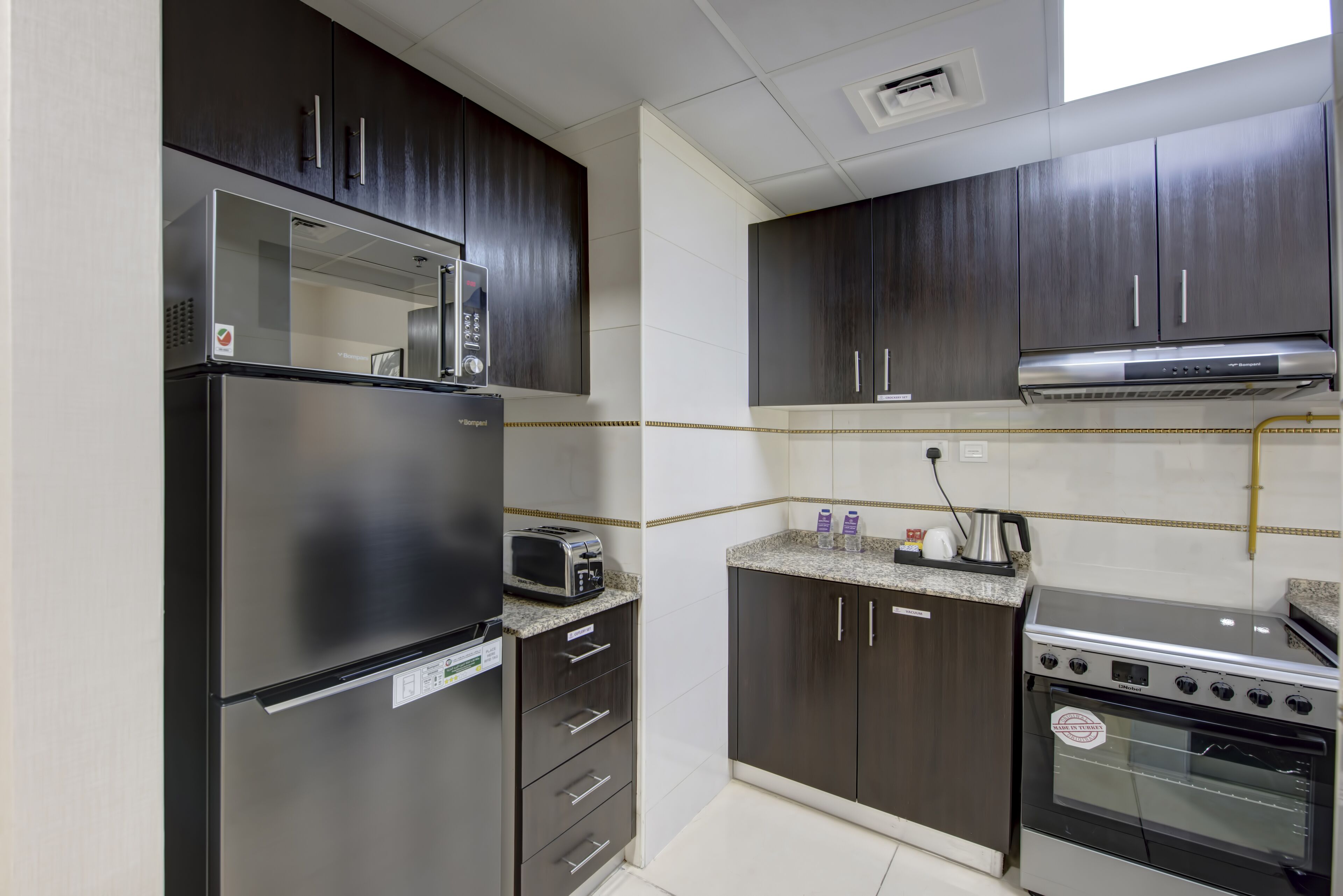Une cuisine de taille réduite avec des meubles de couleur sombre, un réfrigérateur en acier inoxydable, un four à micro-ondes, un grille-pain, une cuisinière et un évier.
