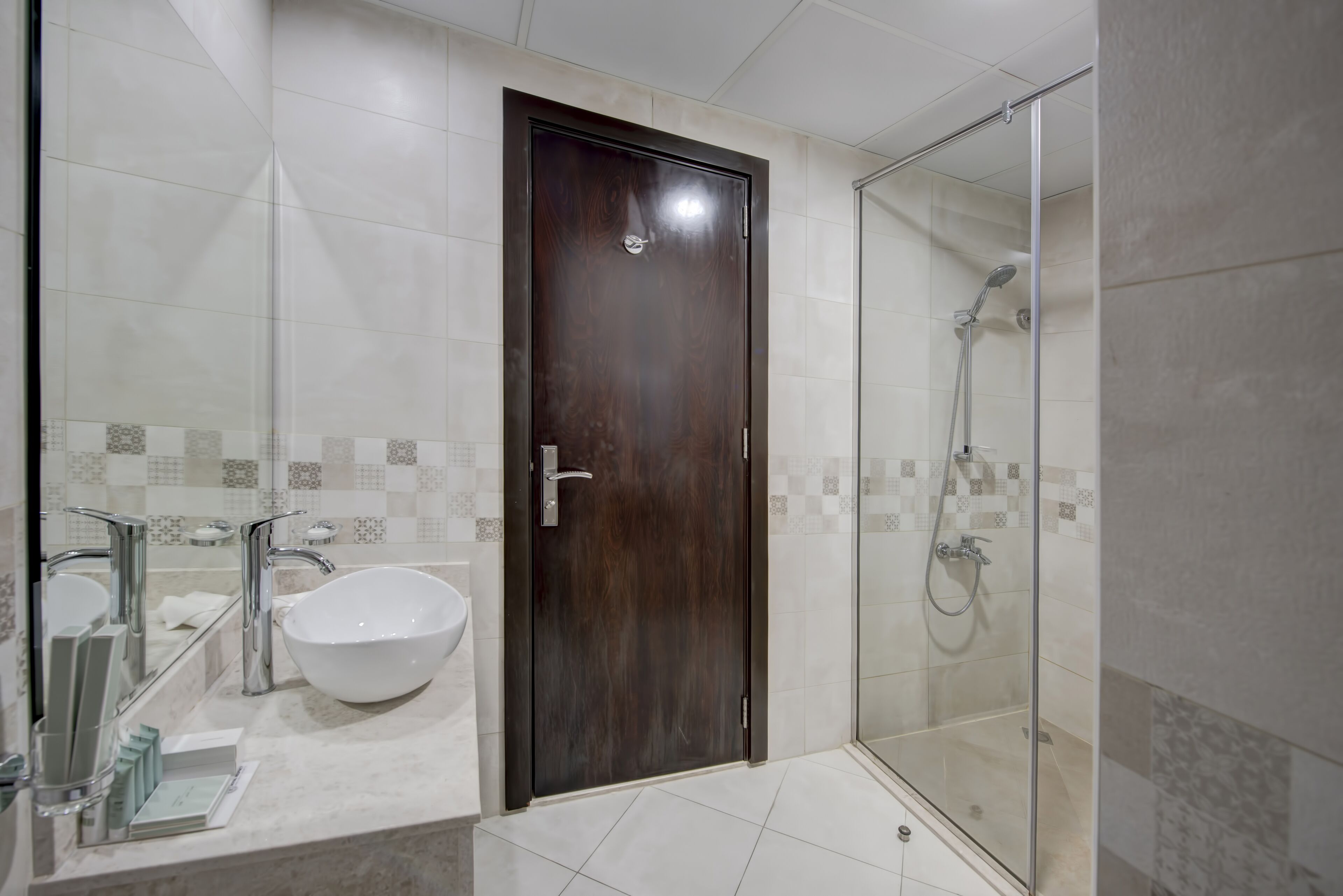 A modern bathroom with a large mirror, a white sink, a dark wooden door, and a glass-enclosed shower area.
