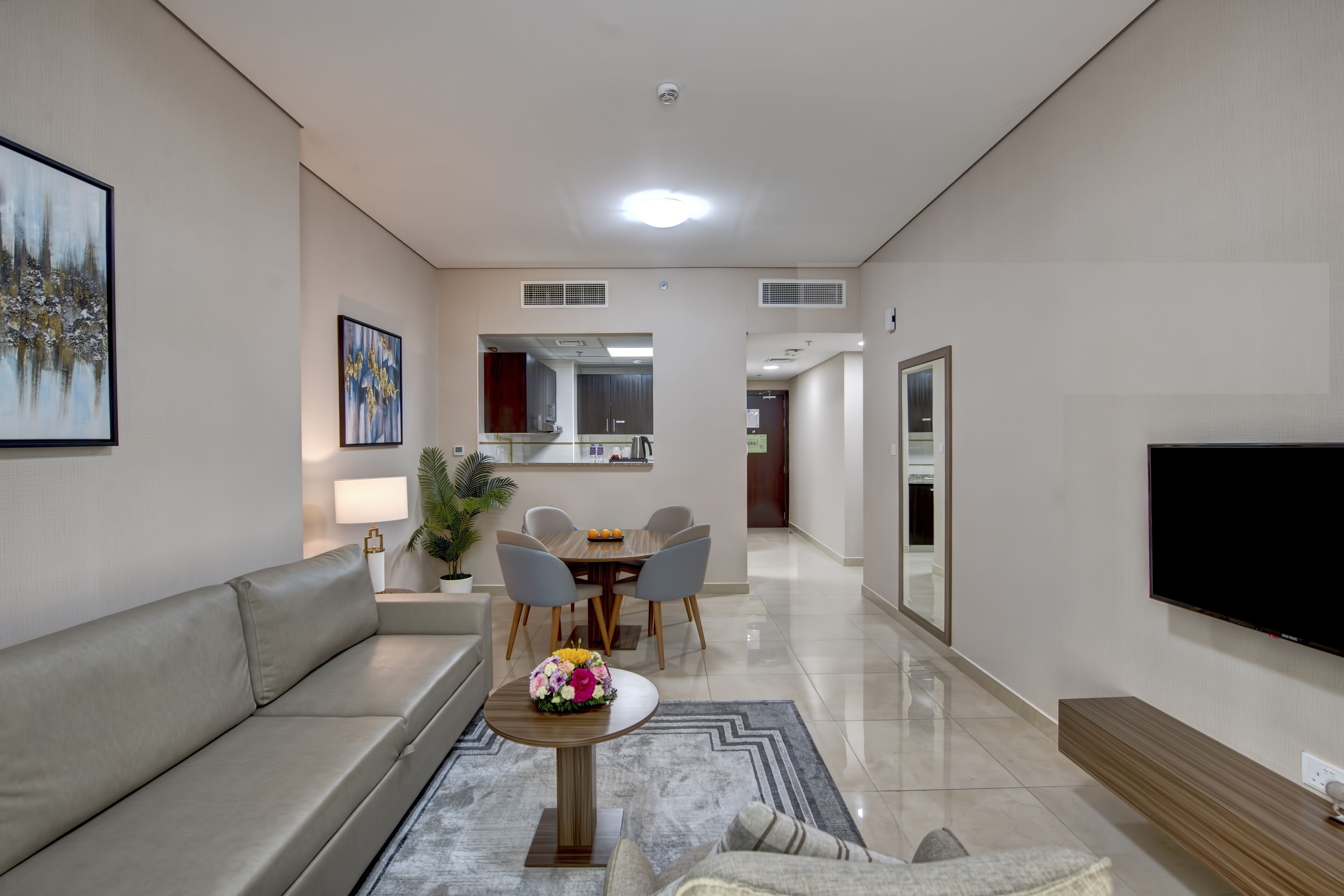 Una sala de estar moderna con un sofá, una mesa de centro con un centro de mesa floral, un comedor con mesa y sillas, un televisor montado en la pared y una cocina al fondo.