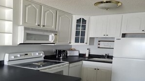 A kitchen with white cabinets, a black countertop, a microwave, a stove, a refrigerator, and a window with frosted glass. - Steps to the Beach! Just “Wait-N-Sea” what this condo has to offer! (North Topsail Beach)