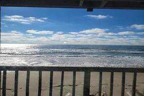 Une vue sur la plage depuis un balcon doté d’une balustrade, avec les vagues de l’océan et un ciel dégagé.