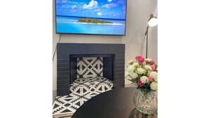 Een moderne woonkamer met een aan de muur bevestigde televisie waarop 'MOVING ART' wordt getoond, een open haard en een vloerkleed met een geometrisch patroon, plus een vaas met bloemen op een bijzettafeltje.