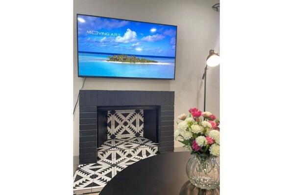 Een moderne woonkamer met een aan de muur bevestigde televisie waarop 'MOVING ART' wordt getoond, een open haard en een vloerkleed met een geometrisch patroon, plus een vaas met bloemen op een bijzettafeltje.